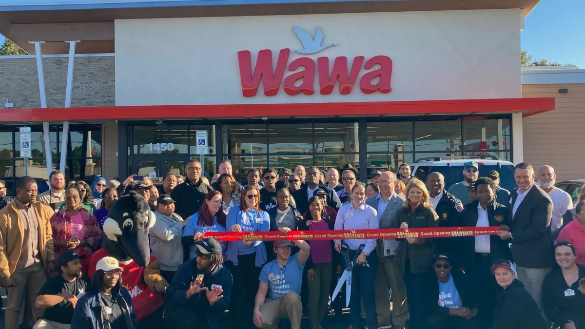 A photo of a number of people and one person in a goose costume standing in front of a store holding a ribbon. The store has a sign that says "Wawa" and the ribbon says "Wawa grand opening."