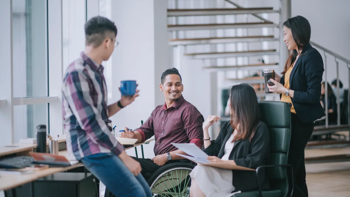 Indian white collar male worker in wheelchair having cheerful discussion leading conversation with colleague in creative office workstation beside window