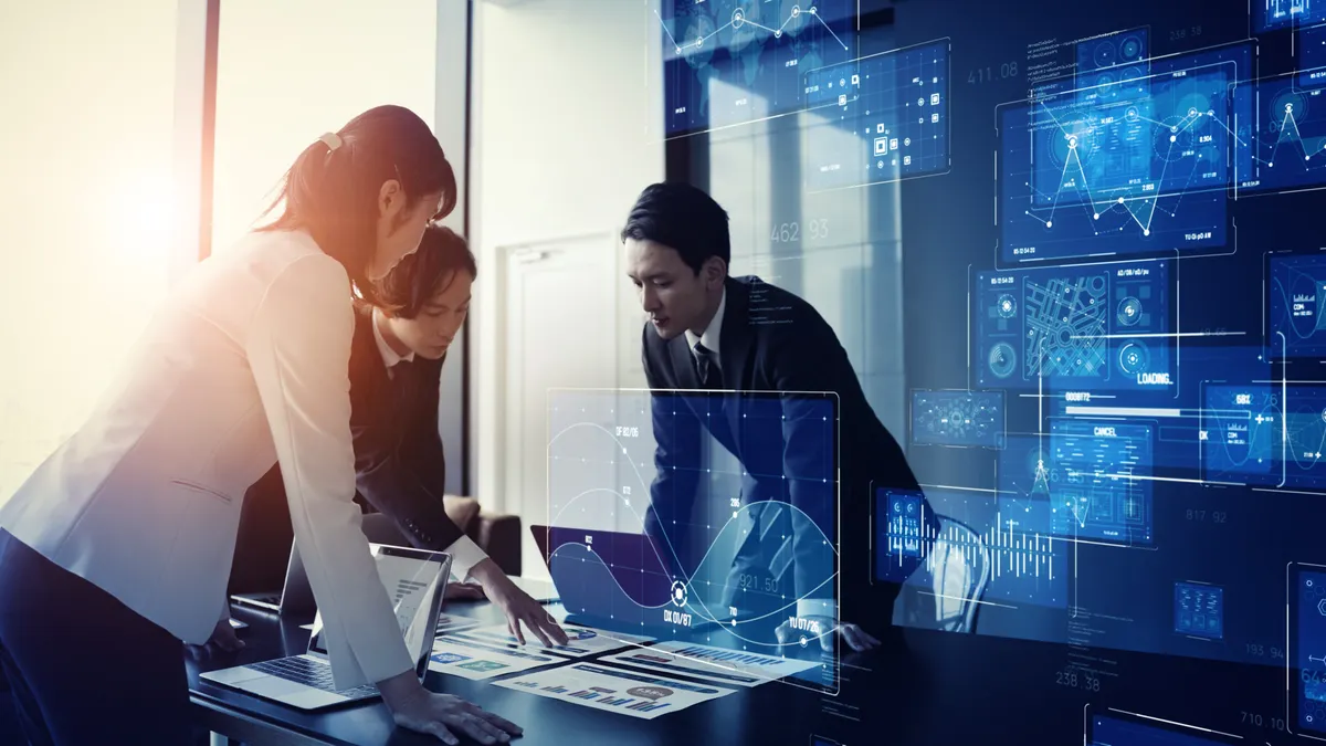 Three people are standing around a table looking at data with digital data on wall.