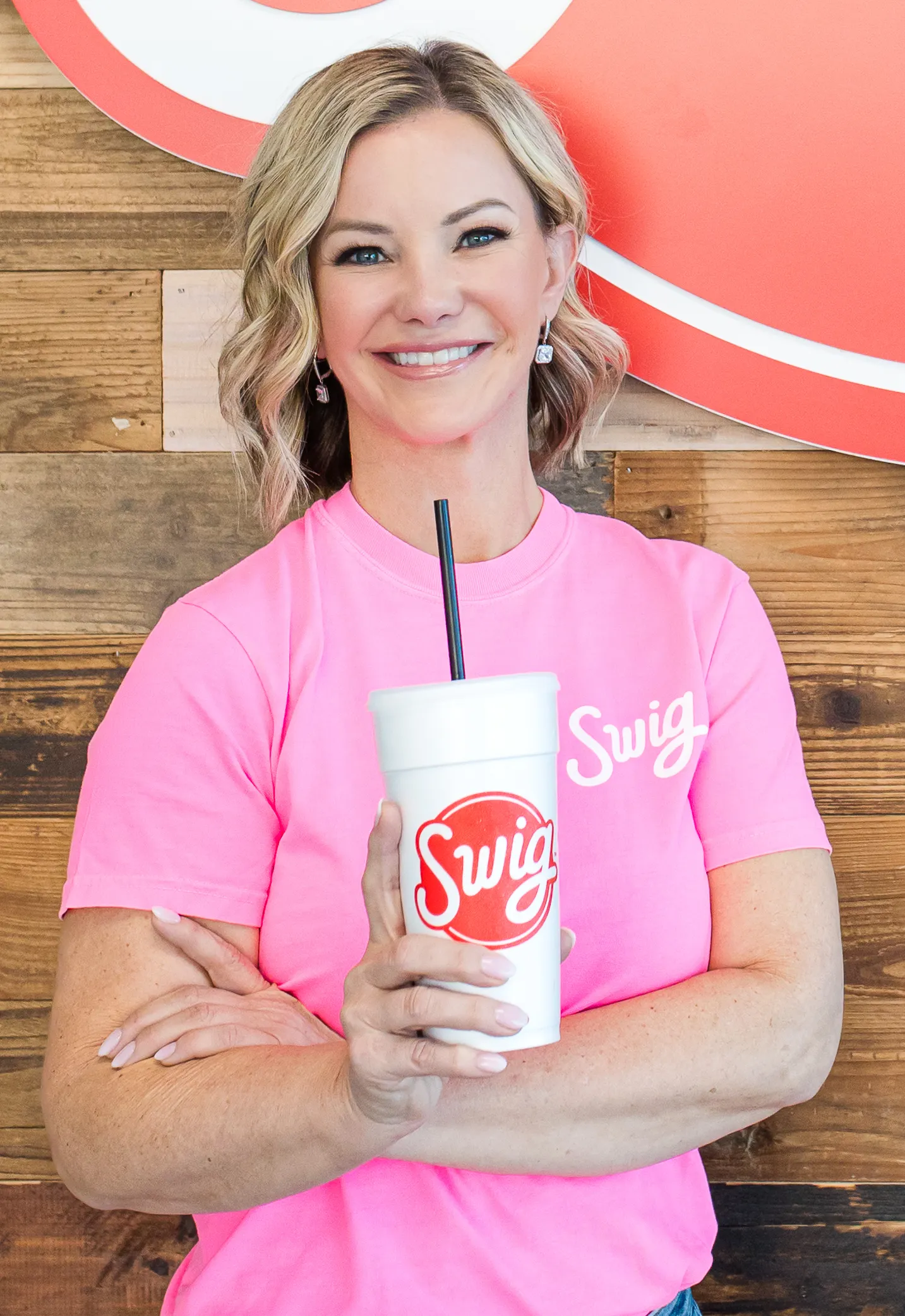 A person wears a pink shirt with a Swig logo and crosses their arms. The person is holding up a white cut with Swig logo.