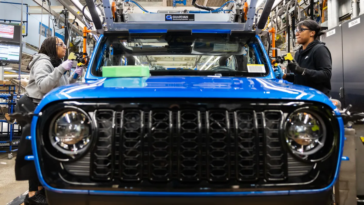 Two workers at Stellantis's Toledo, Ohio Assembly Complex installing the windshield on a blue Jeep Gladiator.