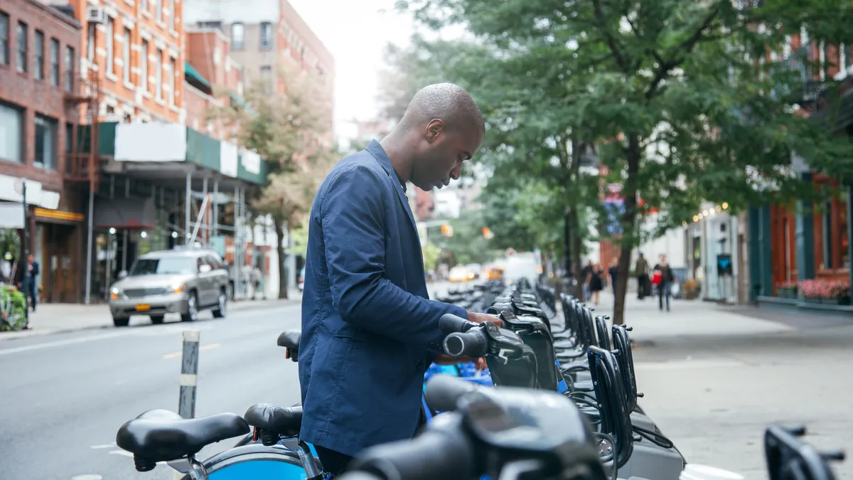 A casually dressed business man in New York City renting a bike from a line of shared bikes on a city street.