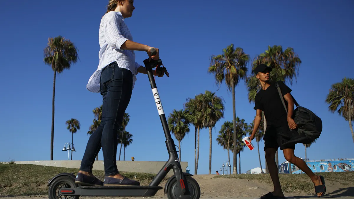 A person rides an e-scooter on a sidewalk in front of palm trees.