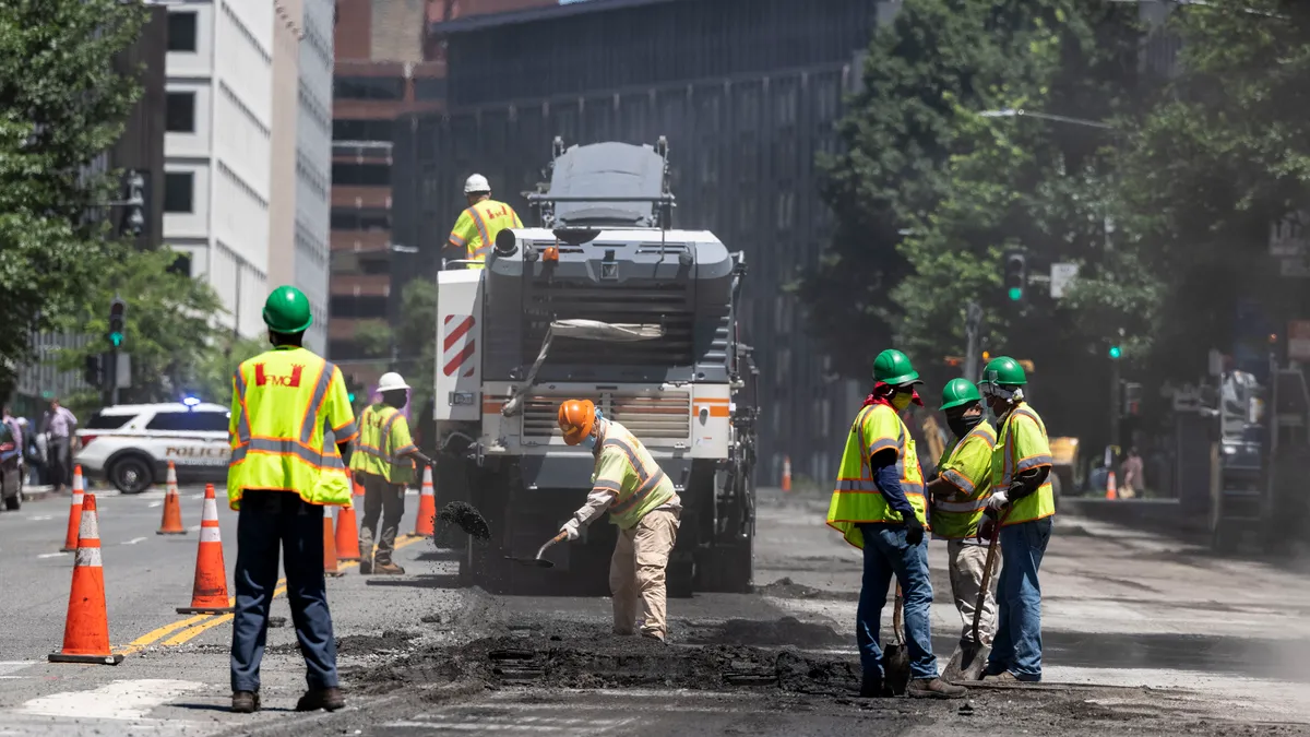 Construction workers in reflective gear shovel asphalt near a large piece of machinery.