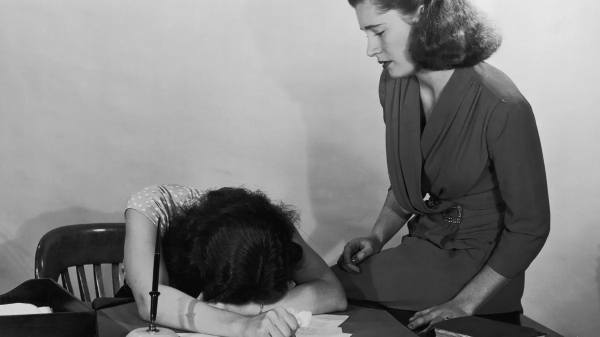 A woman moves to comfort a co-worker who is slumped over her desk in despair; the image, from circa 1940, is in black and white.