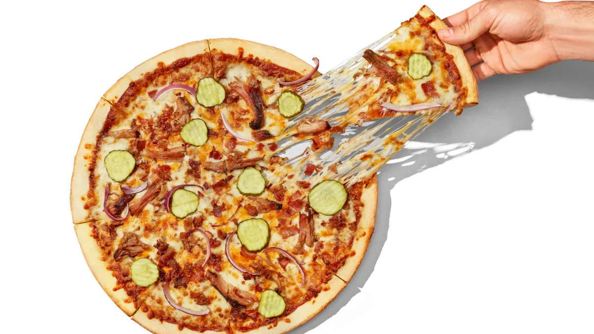 A photo of a pizza with shredded pork, bacon, onions and pickles against a white background. A hand is pulling one slice away from the pizza.