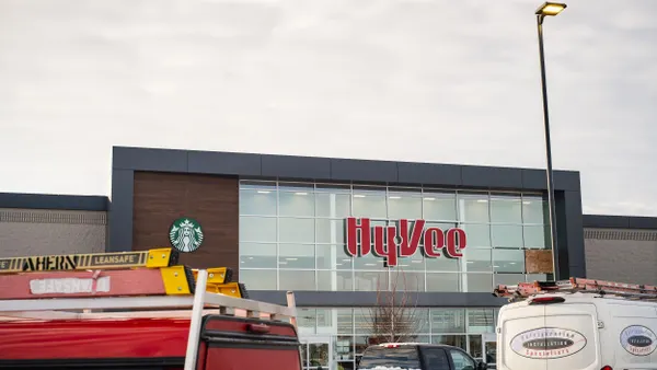 Exterior of a Hy-Vee grocery store with large windows, a Starbucks logo, and red branding, with trucks and vans parked in front of the entrance.