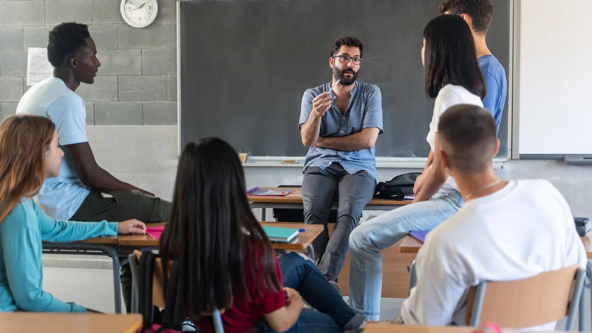 A young bearded teacher is seated on a desk and talking with a diverse group of teenage students seated in a circle in a classroom.