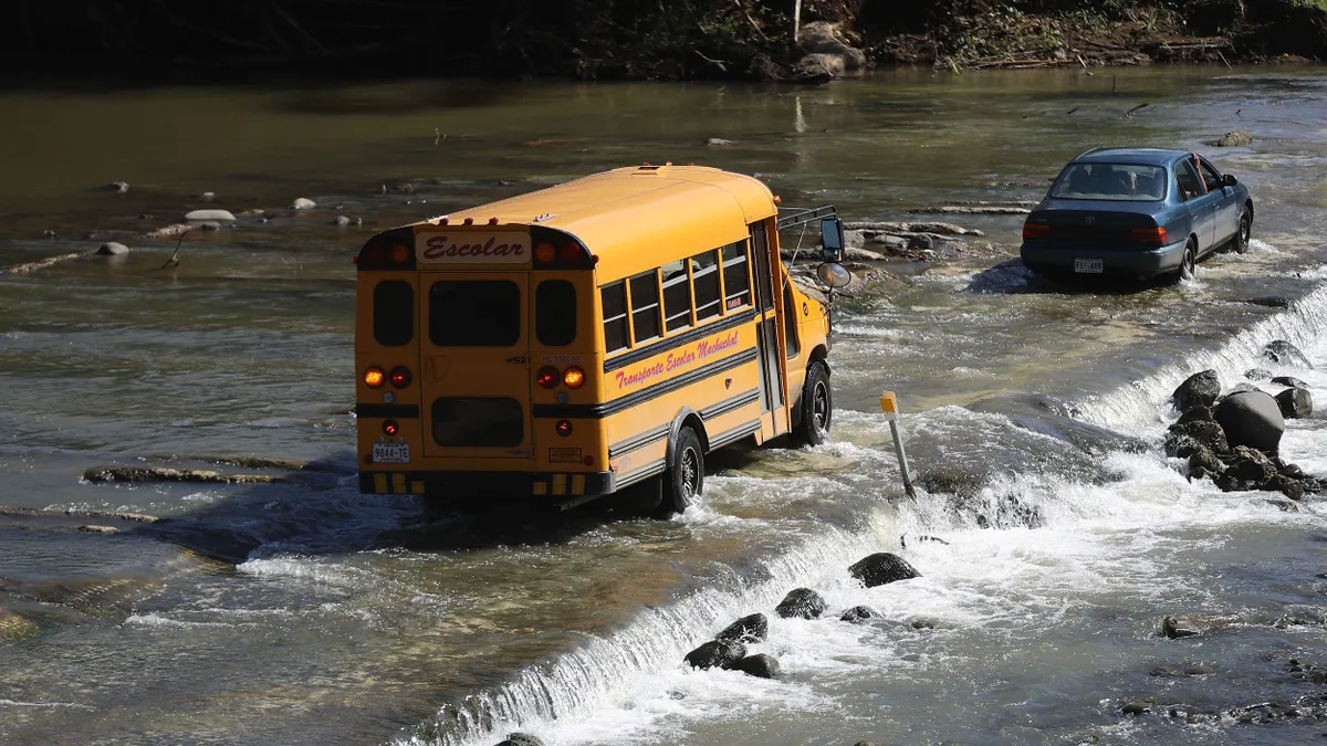 A school bus following a car crosses over a bridge that is covered with water. The photo is taken from up high looking down on the bus, car and bridge.