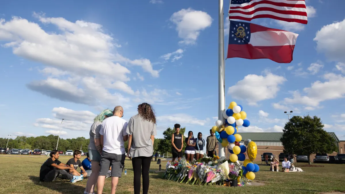 People are standing and sitting around a flag pole in a grass yard. Balloons and flowers are near the flag pole