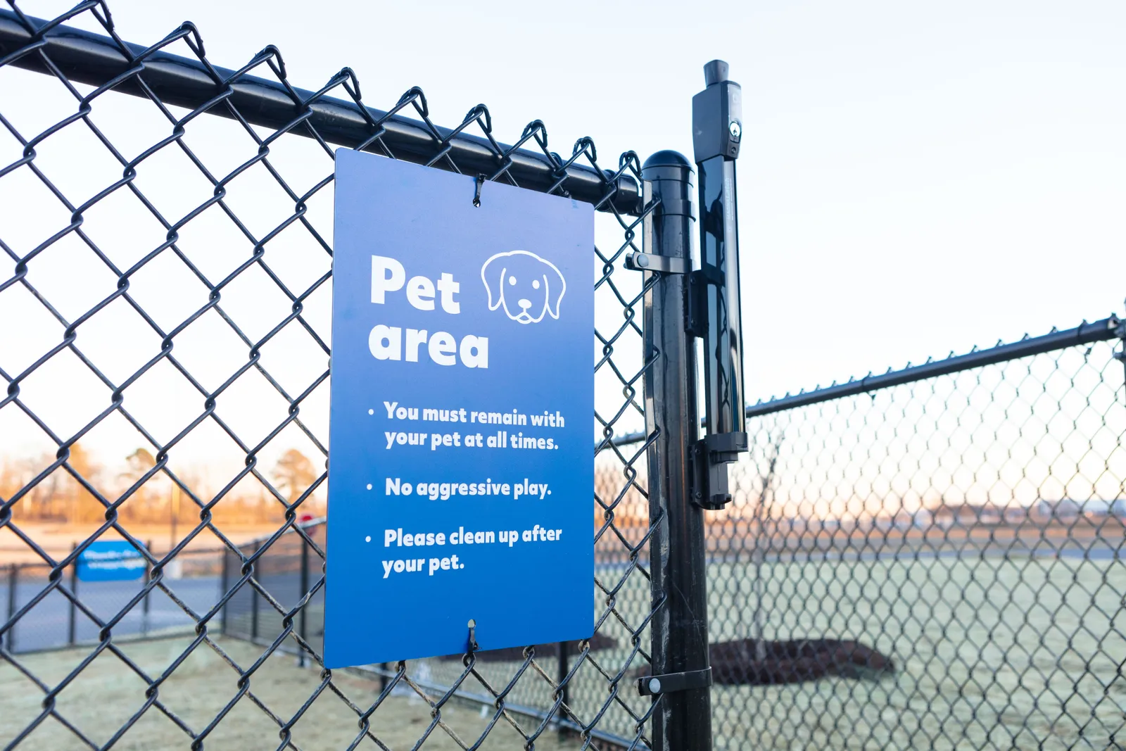 A photo of a chain link fence with a blue sign on it that reads, &quot;Pet area. You must remain with your pet at all times. No aggressive play. Please clean up after your pet.&quot;