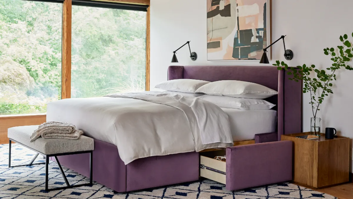 Interior of a bedroom with a bed in the center, rug on the carpet. Two lights are above the headboard on the wall, along with an abstract painting.