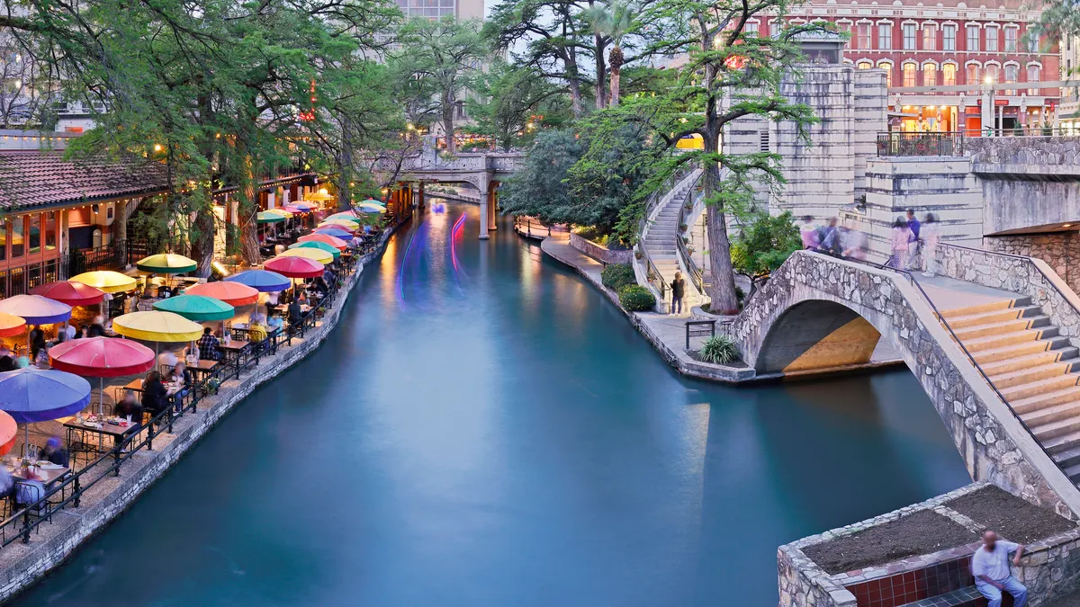 San Antonio's river walk area is shown, with the river in the center of the image, stone stairs on the right and a patio with colorful umbrellas on the left.