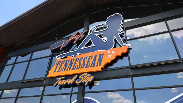 A photo of the exterior of a store, showing a bit of the roof and a wall of windows. A sign on the wall shows the outline of a woman and text saying Dolly's Tennessean Travel Stop.