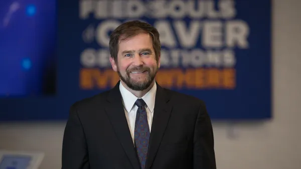 A person stands in front of a blurred background. The person is wearing a blue suit.