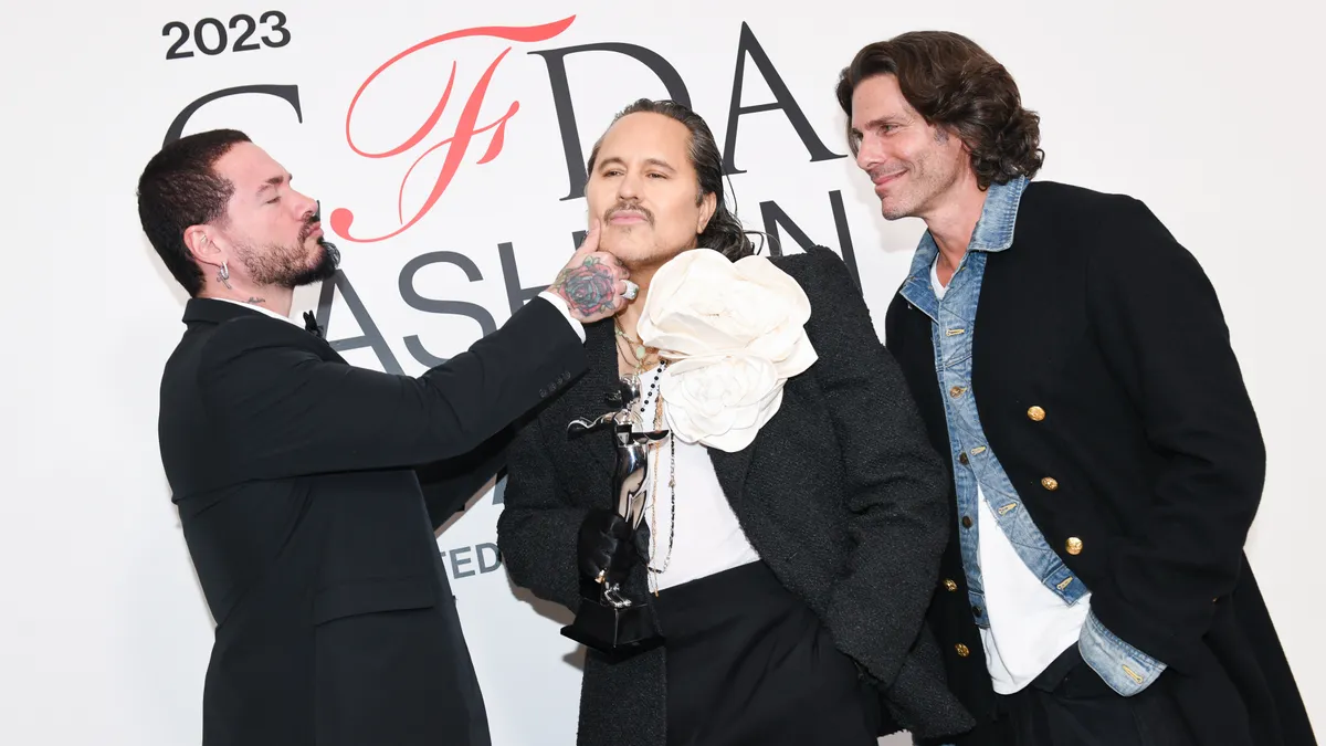 Designer Willy Chavarria wears a large white flower brooch and a black suit. He stands between musician J Balvin and designer Greg Lauren at the CFDA awards show.