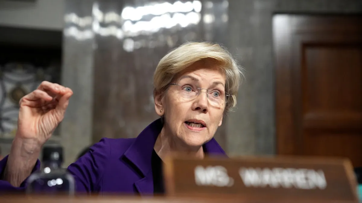 Senator Elizabeth Warren speaks during a legislative hearing, holding her handout to the side. A sign in front of her reads "Ms. Warren."