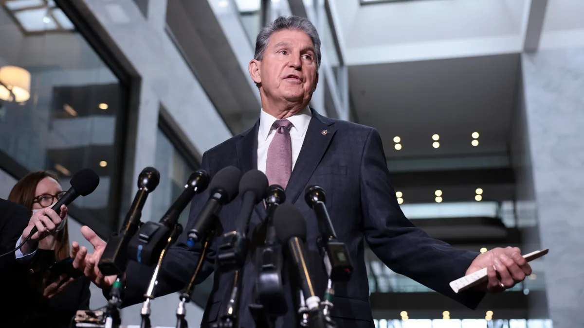 West Virginia Senator Joe Manchin stands behind a row of microphones, with a woman to his right holding another microphone, in an office building.