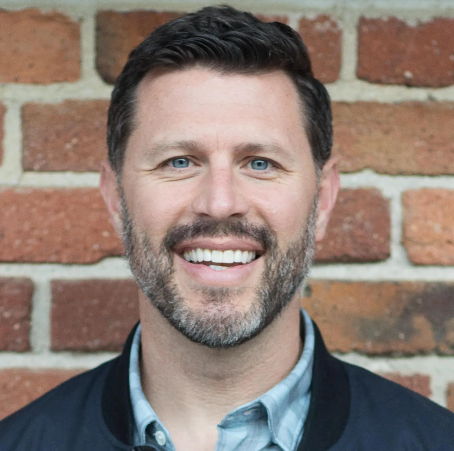 A corporate headshot shows a man in a casual checked shirt smiling at the camera. He has a trimmed beard and mustache.