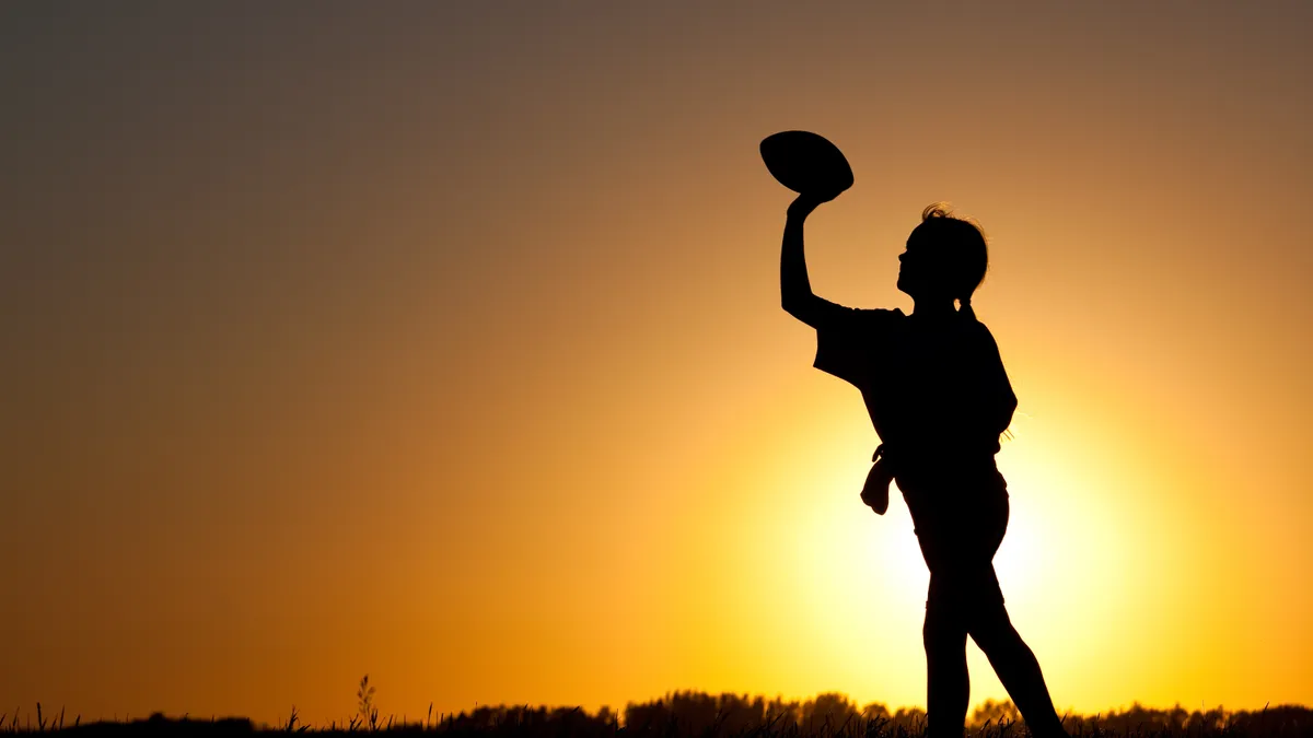 A silhouette of a girl playing football. The silhouette is against an organge background and the girl's arm is up and holding a football.