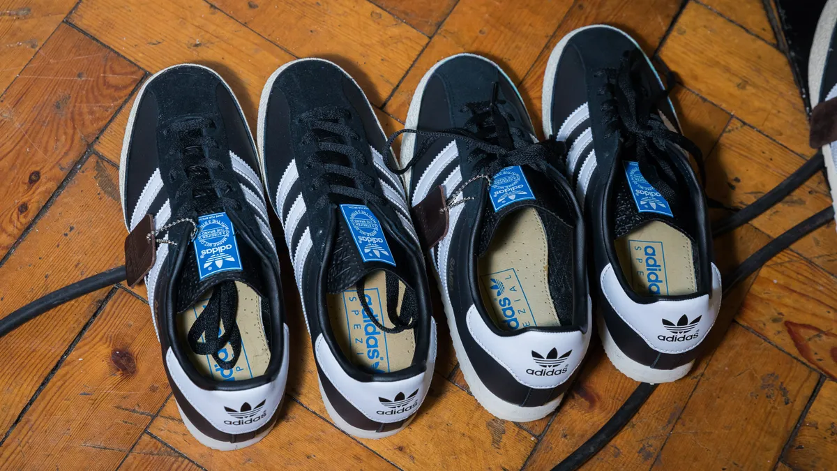 Two pairs of black and white Adidas Sambas sneakers sit side by side unlaced on a wood floor.