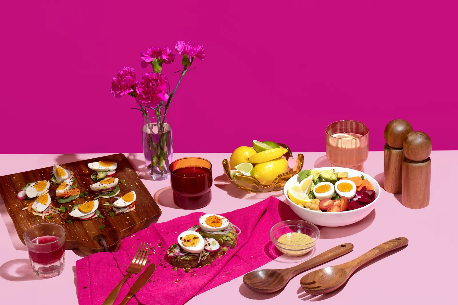 A pink table with a cutting board with wedges of WunderEggs garnished with vegetables, a magenta cloth with WunderEggs on toast and a salad with WunderEggs.