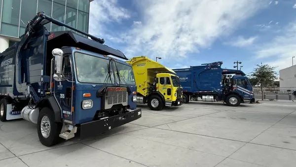 Three front-loading garbage trucks with different branding are all parked on concrete outside a convention center.