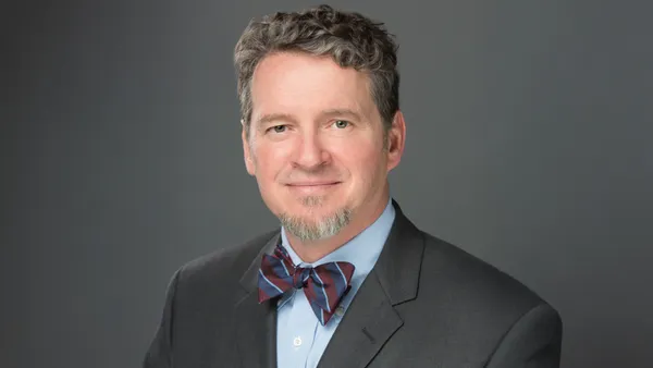 HCA Healthcare's Dr. Thomas Payne wears a gray suit jacket and blue and burgundy striped bow tie, against a gray background.