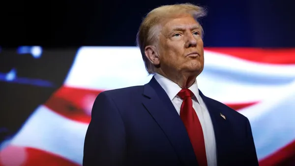 Donald Trump in business attire stands in a stage with an U.S. flag in the background