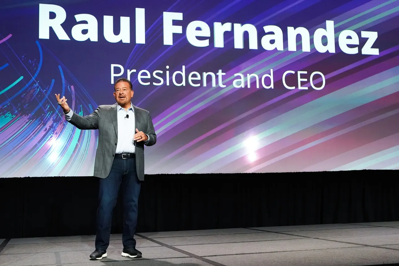 A person dressed in business attire speaks on a stage, behind them is a large screen that reads, "Raul Fernandez President and CEO."