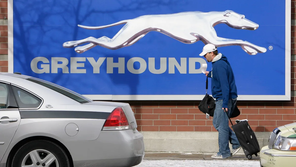 A man carrying luggage walks in front of a building with a large sign that reads "Greyhound."