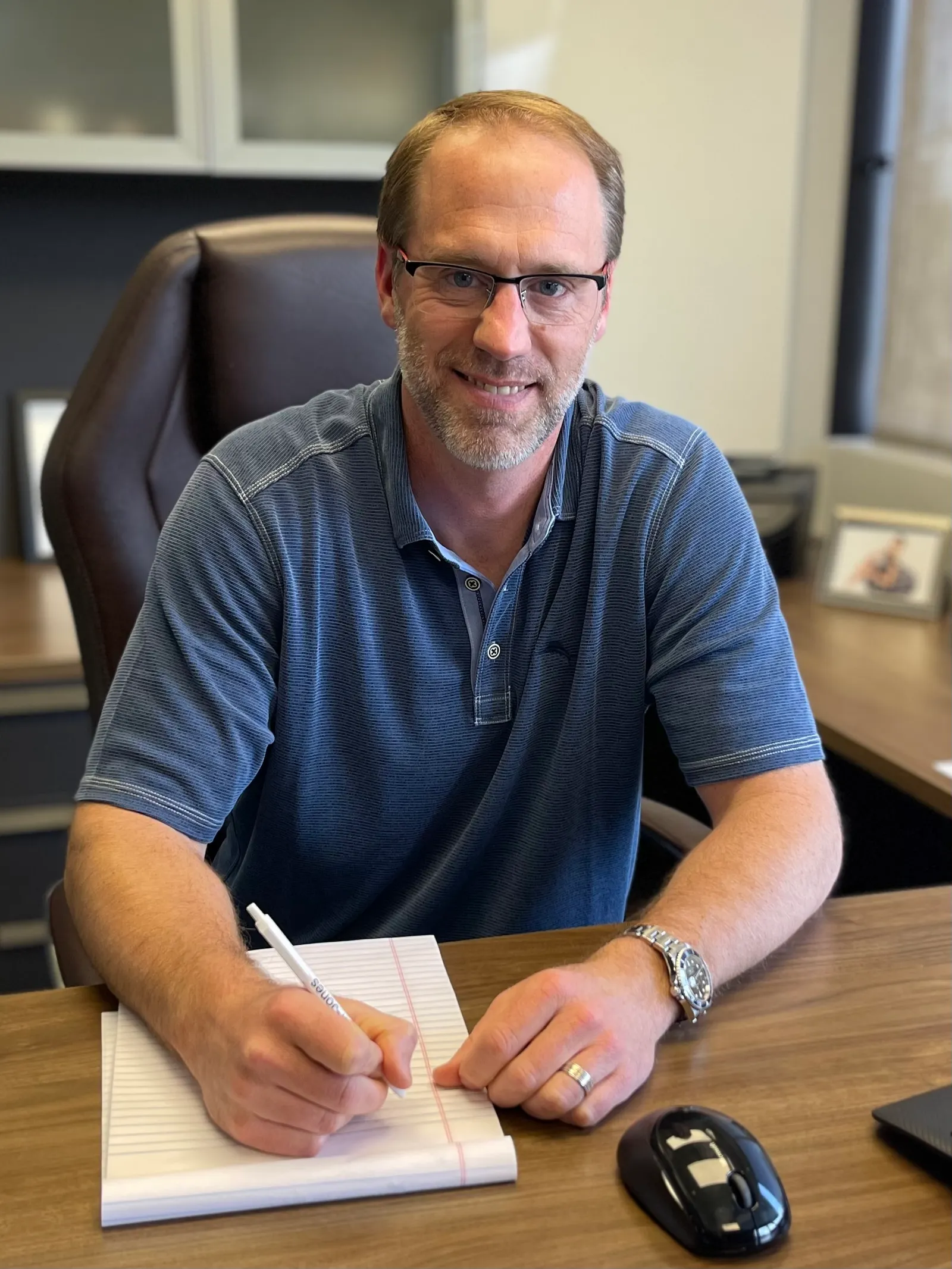 A photo of a person in a blue shirt sitting at a desk, pen in hand, poised to write on a notepad sitting on the desk. They are smiling at the camera.