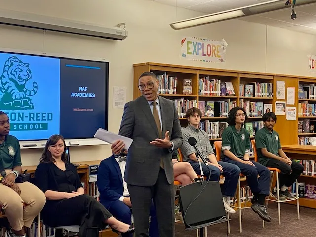 An adult stands in front of a microphone in a school library. Behind the adult are students and educators who are seated.