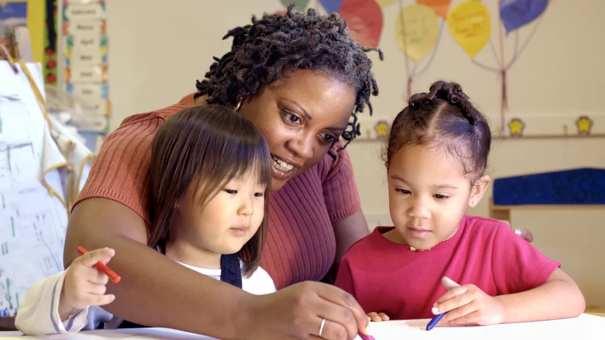 An adult sits at a table with two young students. On the table are Crayons and paper and they are coloring.