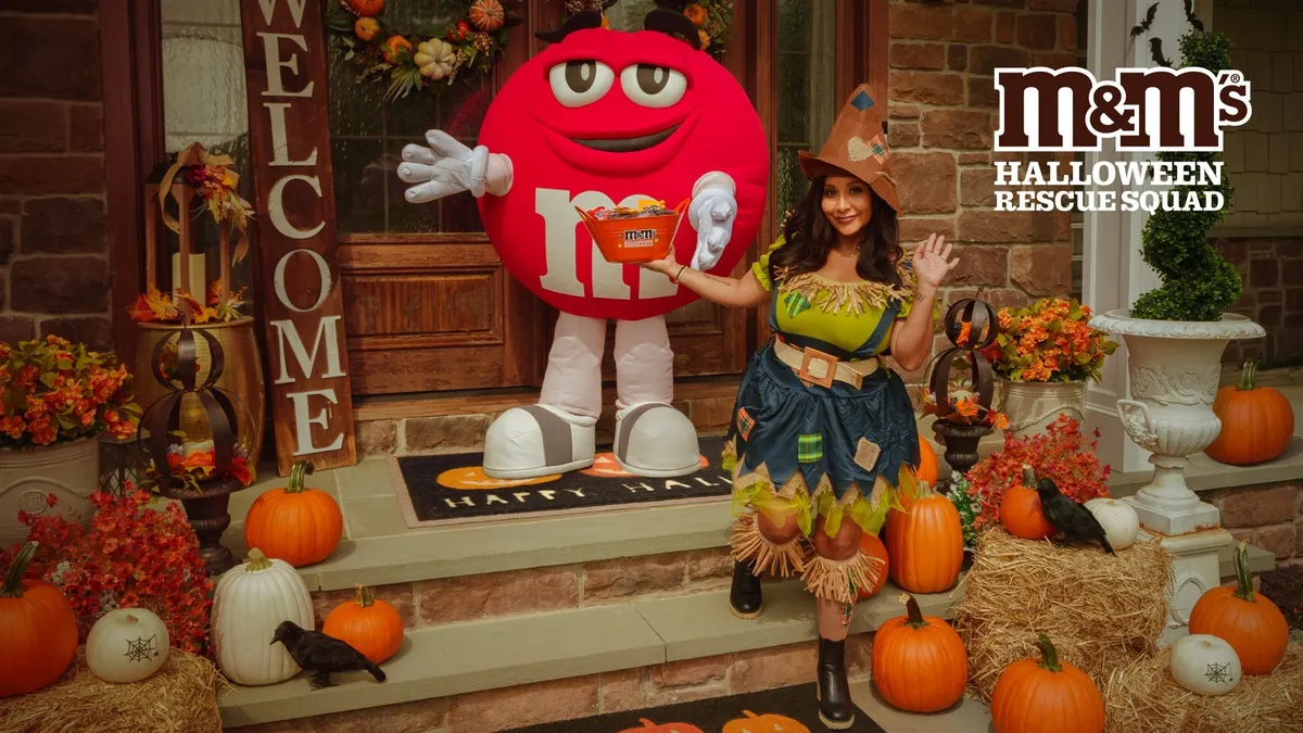 Snooki, dressed as a scarecrow, stands on a decorated front stoop with someone in a red M&Ms costume.