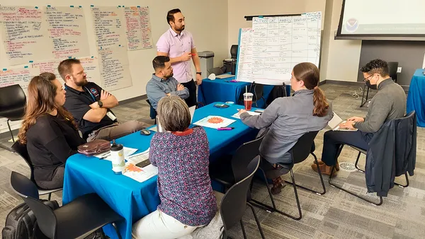 People sit with laptops, pens and paper around a table in a room. On the walls are posters with words written on them in different colored markers.