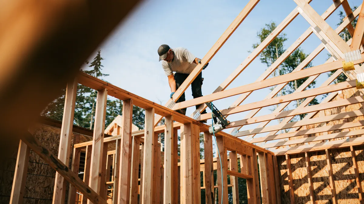 A person in a hat and gloves stands on the wooden beams of home under construction. The person holds a wooden beam.