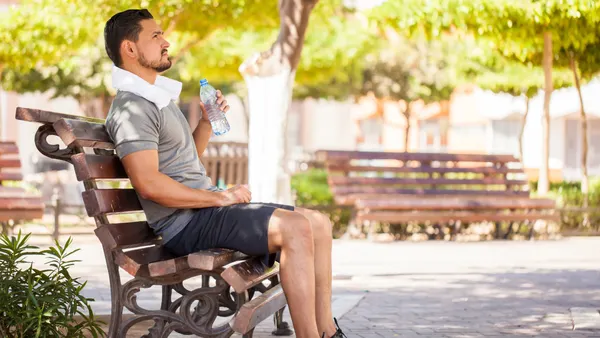 A person wearing shorts and a t-shirt sits on a park bench in the shade of a tree drinking from a plastic water bottle.