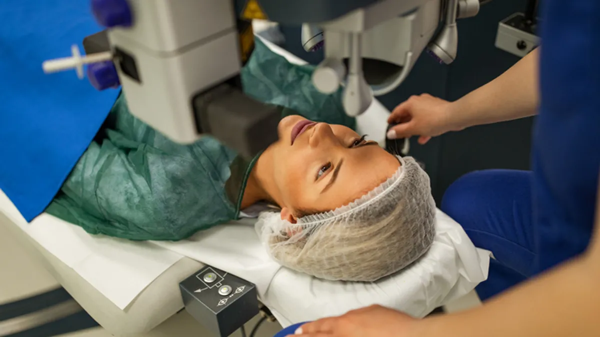 A patient wearing a medical gown and cap sits below a machine for laser eye surgery.