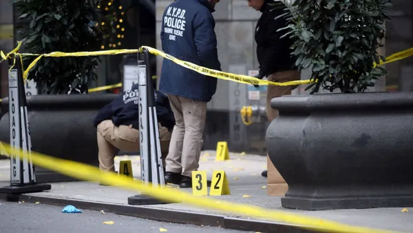 Three police stand on the sidewalk scene of Brian Thompson's killing surrounded by yellow caution tape.