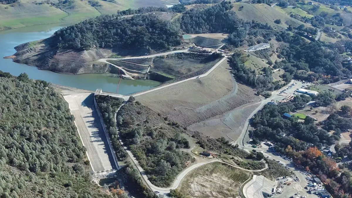 A photo of a dam taken from high in the sky. The dam is surrounded by trees and clear water on one side.