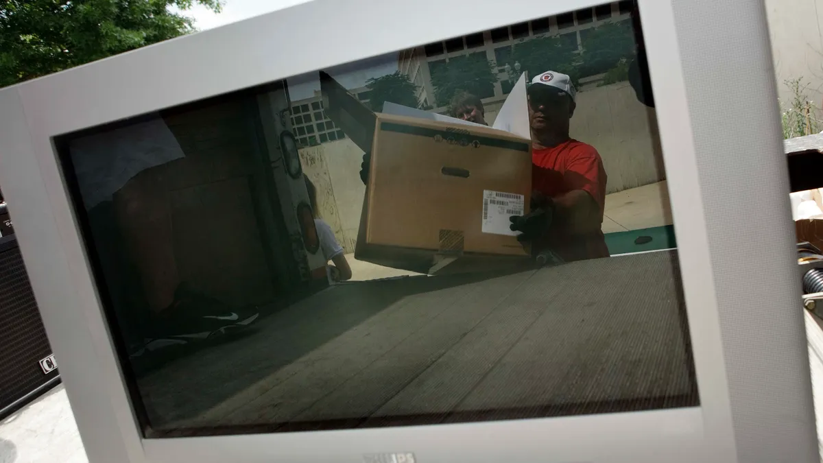 A reflection in a broken television screen shows a man carrying a box