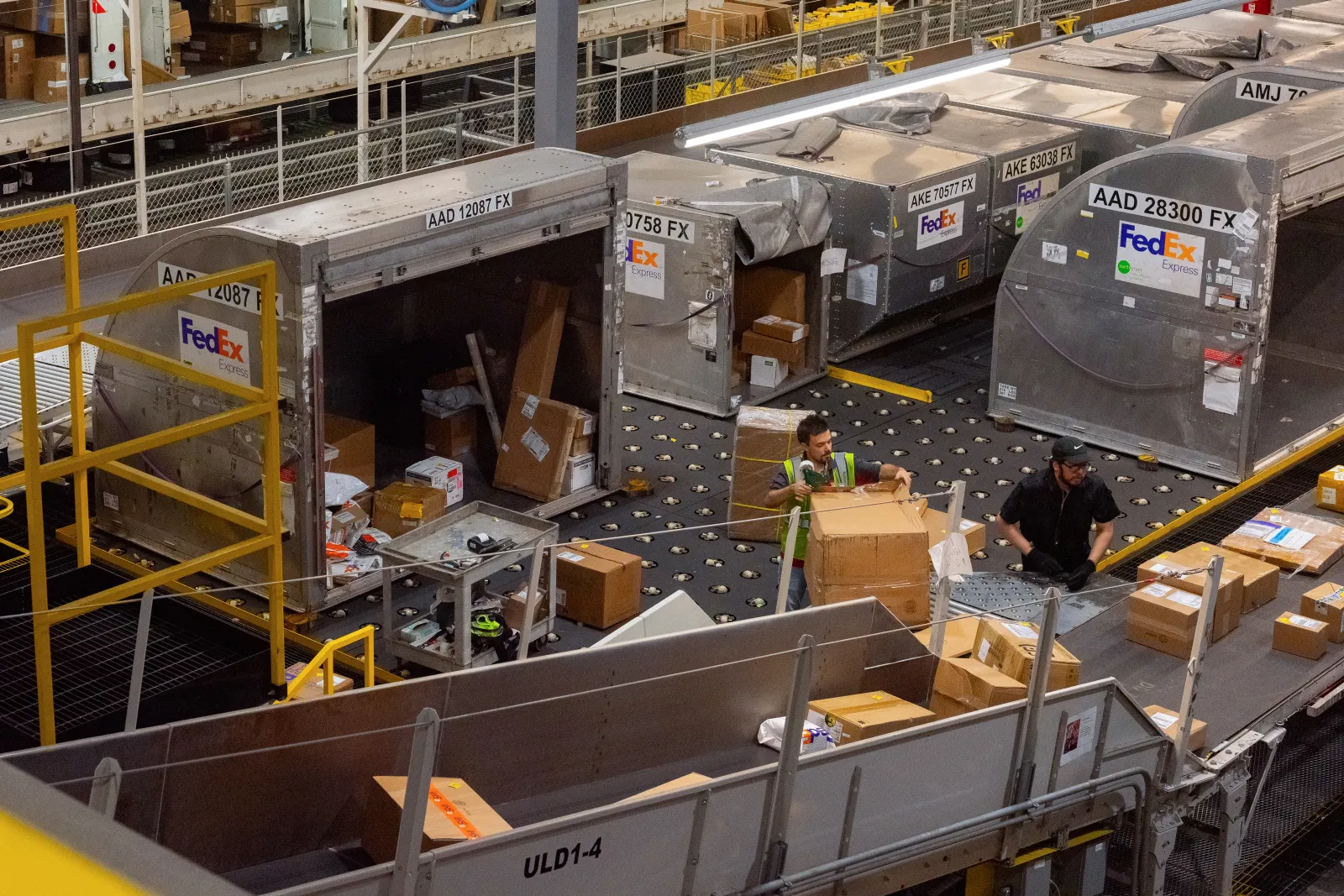 Two employees work in a FedEx warehouse, sorting cardboard boxes from a conveyor belt. They are surrounded by large, metal FedEx Express air freight containers with labels such as "AAD 12087 FX" and "AKE 70577 FX."