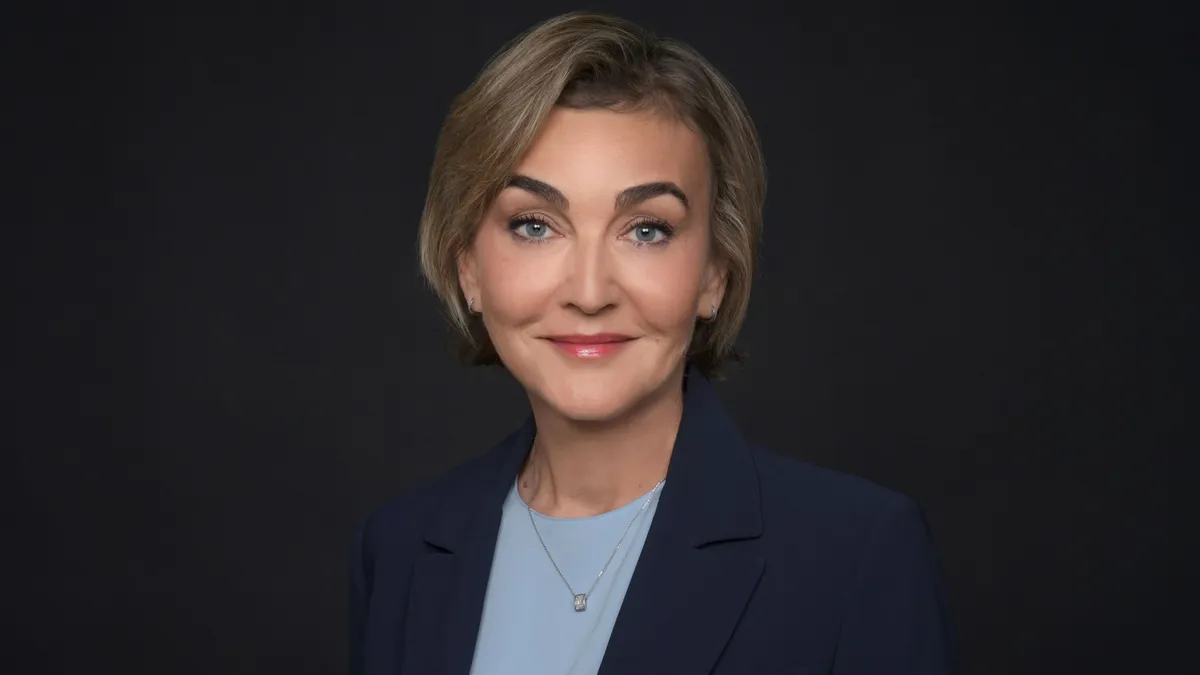 A headshot of a person in front of a blue background. The person is wearing a navy suit.