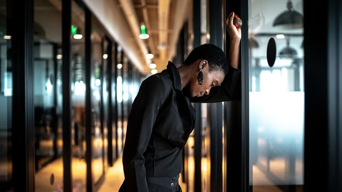 A Black person leans against the frame of a door in their office; their head hangs