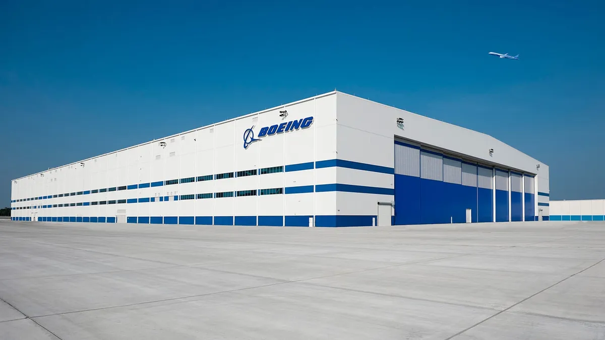 A large warehouse facility, white and blue, stands against a blue sky with the word "Boeing" prominently emblazoned on the side.