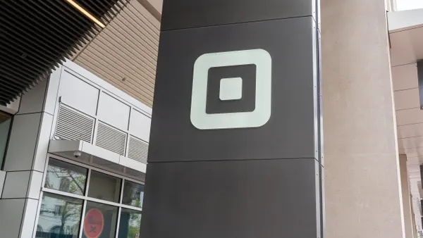 A logo of Square, inc. is displayed on a metal sign tht is positioned infront of a commerical building. 