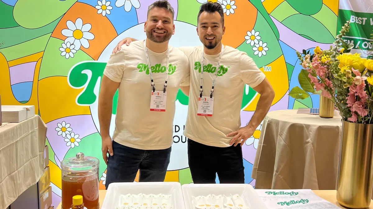 Darko Mandich and Aaron Schaller stand at a colorful booth for the Mellody brand with a branded backdrop and a table in front of them with sample spoons of honey.
