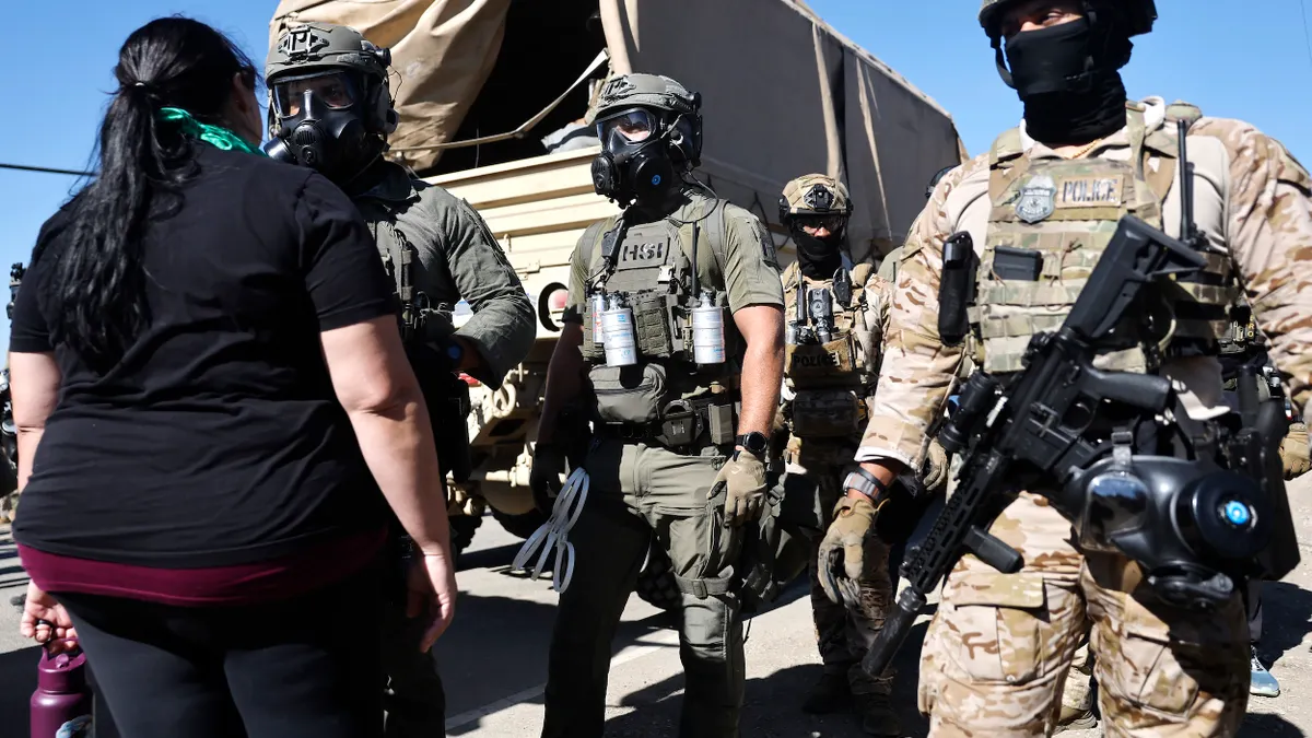People wearing camouflage suits that say POLICE, helmets and black scarves over their faces, carrying guns, facing a person wearing a black t-shirt and black plants.