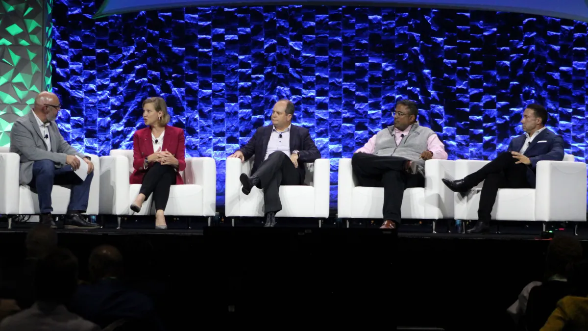 Five people sit in white chairs on a stage. One of them, the second from the left, is speaking.